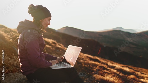 Woman working on laptop in sunset mountains. Young freelancer tourist caucasian female typing on notebook sitting on autumn grass meadow landscape. Concept of remote online work, study or business photo