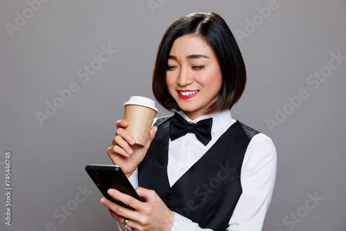 Smiling asian woman dressed in waitress uniform holding coffee paper mug and browsing internet on smartphone. Receptionist enjoying tea and engaging with social media networks on mobiel phone photo