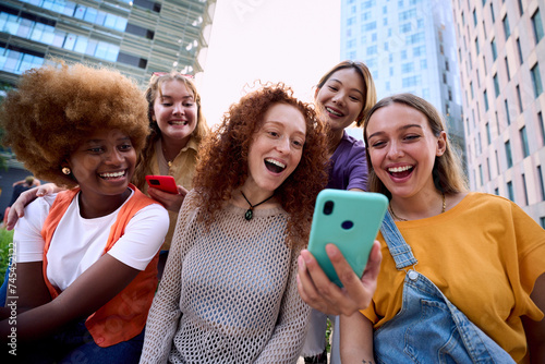Young group of gen z people having fun surprised using cell phone together outside watching something funny at mobile. Cheerful community of student enjoying social media content. Multiethnic friends photo