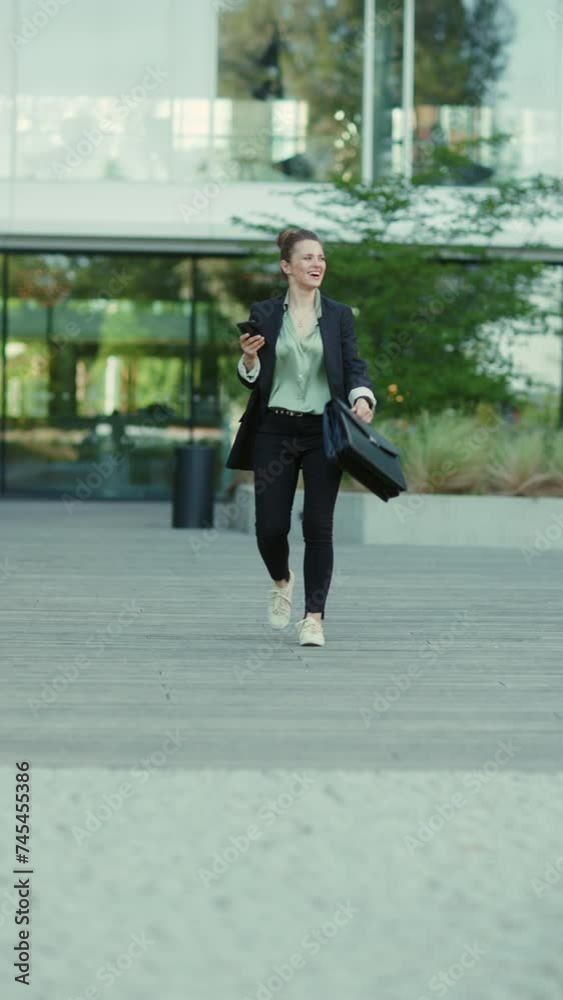 Full length portrait of happy modern 40 years old woman employee near business center in black jacket with smartphone and briefcase walking. Full length portrait of happy modern 40 years old woman employee near business center in black jacket with smartphone and briefcase walking.