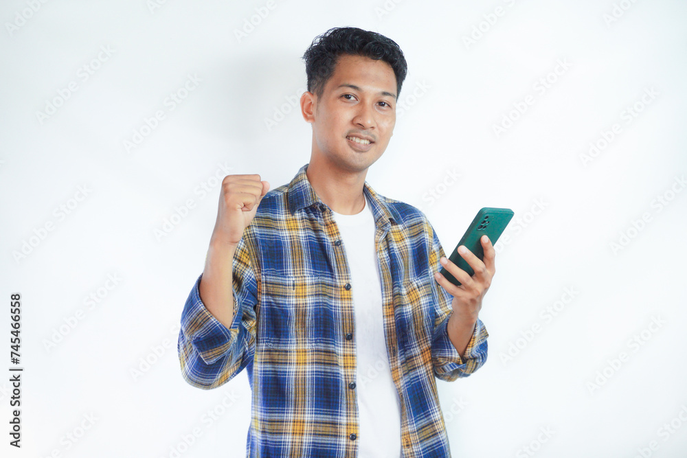 Adult Asian man showing excited expression while clenched fist and holding mobile phone Adult Asian man showing excited expression while clenched fist and holding mobile phone