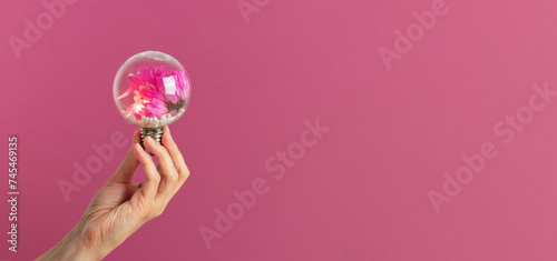 Magenta flowers inside the light bulb in woman's hand against purple background. Concept of natural power, eco-friendly energy, green solutions. © elcovalana