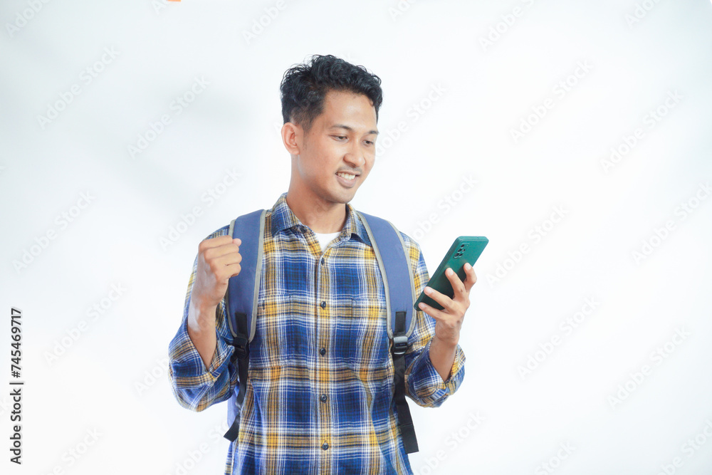 Excited young Asian man student in casual clothes backpack holding mobile cell phone and saying yes, celebrating success isolated on white background. high school university college concept Excited young Asian man student in casual clothes backpack holding mobile cell phone and saying yes, celebrating success isolated on white background. high school university college concept