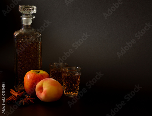 Apple-peach liqueur with cinnamon and anise on a black background, homemade alcohol in a decanter and glasses with spices and ripe fruits.