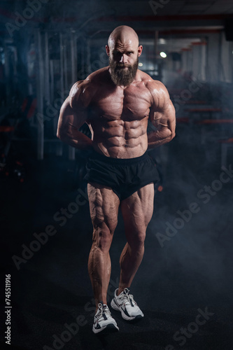 Muscular bald man posing in shorts. Bodybuilder showing off his shape in the gym. 