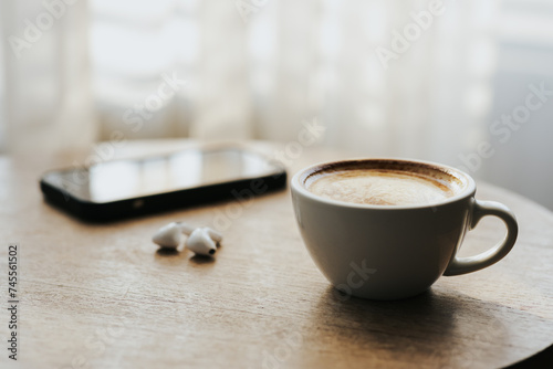 A cup of hot latte and a cell phone, headphones on a sunny wooden table by a window with white curtains. Vintage tones. Free space to create a message. photo