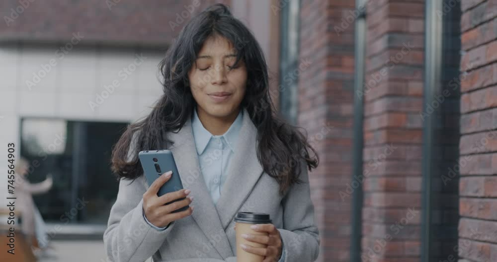 Dolly shot of confident Middle Eastern businesswoman walking in street drinking to go coffee and using smartphone. Modern lifestyle and businessperson concept.
