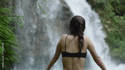 Beautiful woman in black bikin top stares out to gorgeous tropical waterfall in forest