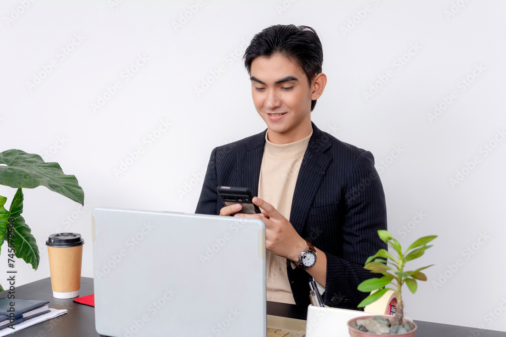 Young Asian businessman smiling or amused while checking his cellphone, with a coffee cup and computer on his desk. Young Asian businessman smiling or amused while checking his cellphone, with a coffee cup and computer on his desk.