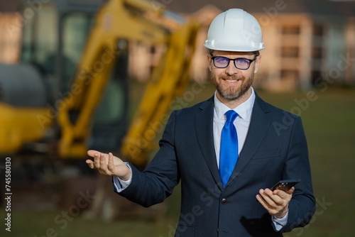 Civil engineer worker at a construction site. Mature engineer worker. Man in suit and hardhat helmet at construction site. Middle aged head civil engineer worker standing outside near excavator.