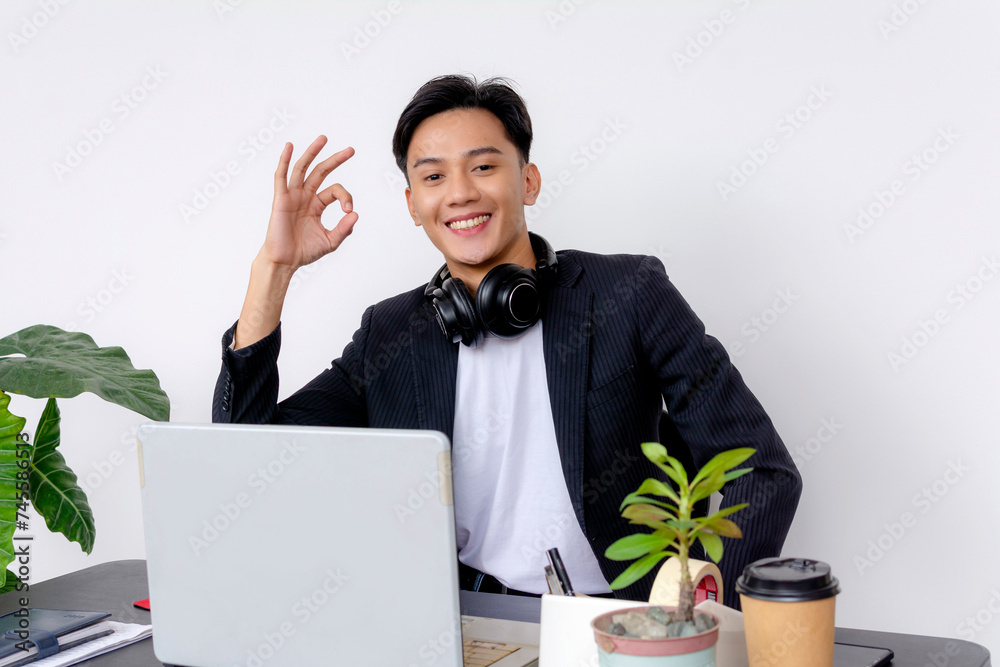 Cheerful young professional making an OK sign while sitting at his work desk with a laptop, headphones, and coffee. Cheerful young professional making an OK sign while sitting at his work desk with a laptop, headphones, and coffee.