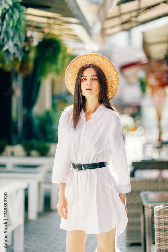 A cute girl in a hat walks along a tourist street among cafes. a young brunette in a white dress enjoys the city