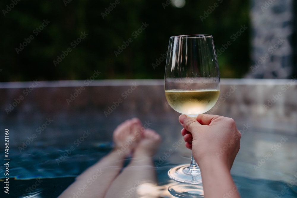 Unrecognizable young woman with feet up relaxing with glass of wine in hot tub outdoor in nature.