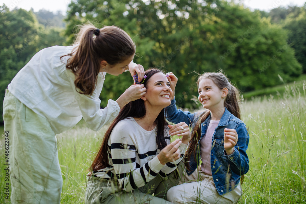 Beautiful mother with daughters, picking flowers, putting in hair, sitting in grass at meadow. Concept of Mother's Day and maternal love. Beautiful mother with daughters, picking flowers, putting in hair, sitting in grass at meadow. Concept of Mother's Day and maternal love.