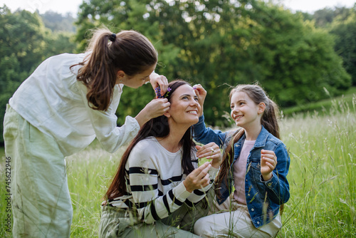 Beautiful mother with daughters, picking flowers, putting in hair, sitting in grass at meadow. Concept of Mother's Day and maternal love.