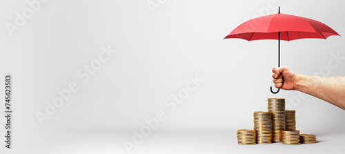 A hand firmly holds a red umbrella, shielding a rising sequence of coin stacks, hinting at financial security