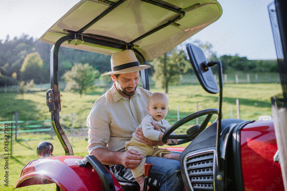 Farmer father riding tractor with his little baby son. Baby growing up on family farm. Concept of multigenerational farming.