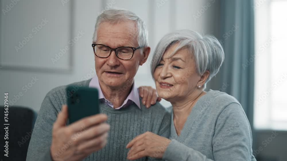 Senior couple having video call at home. Old gray haired man woman chatting talking on smartphone sitting on couch. Happy pensioners enjoying phone conversation with family, friends smiling laughing. Senior couple having video call at home. Old gray haired man woman chatting talking on smartphone sitting on couch. Happy pensioners enjoying phone conversation with family, friends smiling laughing.