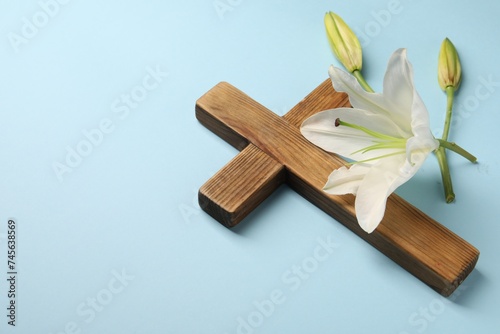 Wooden cross and lily flowers on light blue background, space for text. Easter attributes