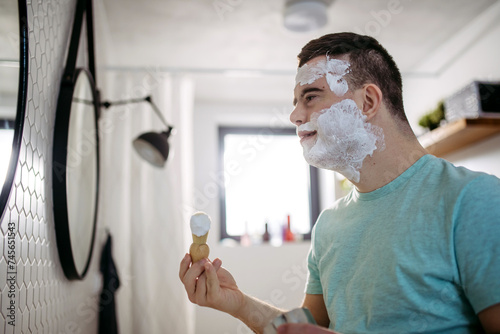 Young man with down syndrome learning how to shave, applying shaving foam all over his face, looking at mirror. photo