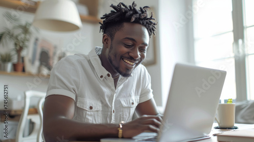 A joyful young man with dreadlocks works on a laptop in a bright, cozy room
