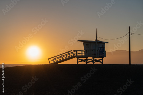 A lifeguard tower stands silhouetted against a colorful sunset on a beach. The sky is ablaze with orange, pink, and purple hues, and the water reflects the vibrant colors © Steeve