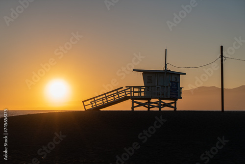 A lifeguard tower stands silhouetted against a colorful sunset on a beach. The sky is ablaze with orange, pink, and purple hues, and the water reflects the vibrant colors