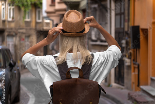 Photo from the back of a young blonde woman with a backpack holding her hat with her hands on a beautiful street in Istanbul,Turkey. Stylish girl walks along an atmospheric city street, travel