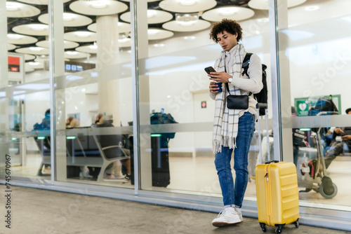 Young man with suitcase using smart phone leaning on glass wall at airport photo
