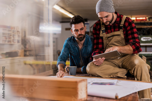 Carpenter sharing tablet PC with colleague sitting on workbench at workshop photo