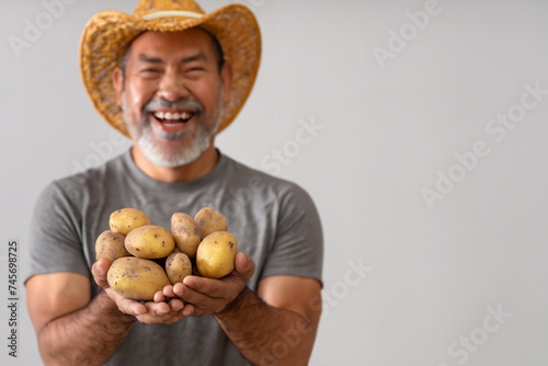 man with potatoes. cheerful asian man in a gray T-shirt and wicker hat stands on a light background with potatoes in his hands, farming concept