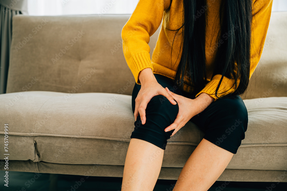 Illustrating Health Care, Close-up of young woman holding painful knee on sofa displaying chronic tendon arthritis. Depicting pain recovery and tendon inflammation. Illustrating Health Care, Close-up of young woman holding painful knee on sofa displaying chronic tendon arthritis. Depicting pain recovery and tendon inflammation.