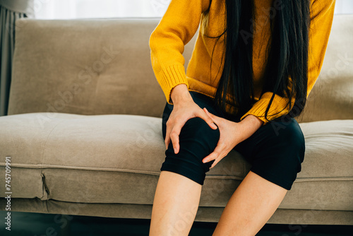 Illustrating Health Care, Close-up of young woman holding painful knee on sofa displaying chronic tendon arthritis. Depicting pain recovery and tendon inflammation.