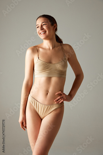 Beautiful, smiling, young brunette girl with well-kept skin, health fit body posing in underwear against grey studio background. Concept of body and health care, female beauty, wellness