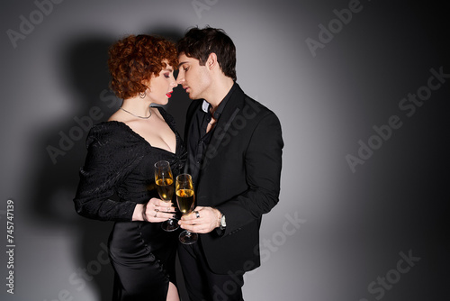 appealing woman in black dress hugging her handsome boyfriend on gray backdrop, champagne in hands