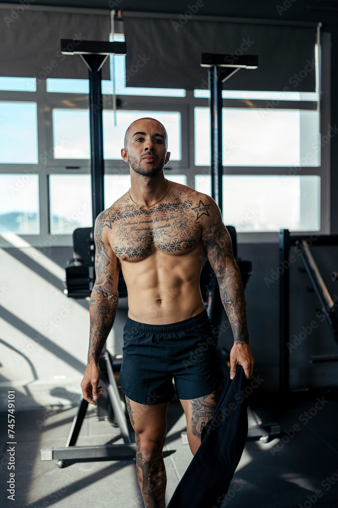 Chico joven tatuado y musculoso posando en gimnasio sin camiseta y con ropa moderna