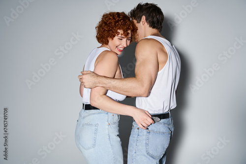 cheerful alluring man and woman in jeans posing on gray backdrop and smiling happily, sexy couple