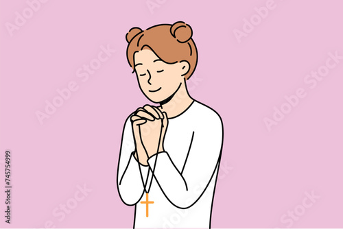 Little girl reads prayer, crossing palms near chest and holding christian cross in hands. Happy pre-teen child praying, feeling involved in catholicism and christian religion at early age