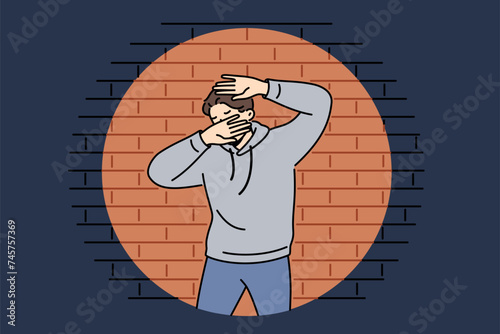 Man criminal in light of police lantern stands near brick wall and covers face with hands. Young guy criminal who committed robbery or murder was cornered after pursuit and investigation