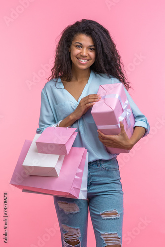 Studio vertical portrait of happy smiling beautiful casually dressed young woman posing standing with gift box and bunch of shopping bags in hands over pastel pink background