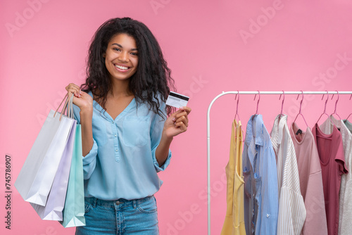 Attractive stylish smiling elegant young woman posing with with shopping bags and credit card in hands while standing near the rack of pastel colored clothes isolated on pink background