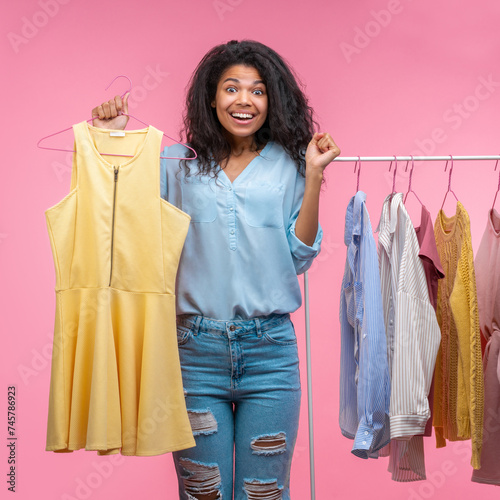 Studio portrait of excited smiling attractive young woman holding hanger with illuminating yellow dress in hand while standing near the rack of pastel clothes in showroom