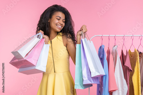 Studio portrait of happy confident girl with charming smile  posing with a bunch of shopping bags in hands, standing near clothing rack, isolated over pastel pink background