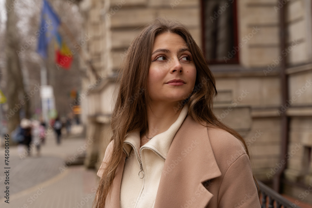 Young beautiful woman on a walk. A girl in a beige coat stands near a classical-style building and looks into the distance. The flag of the European Union is in the background.