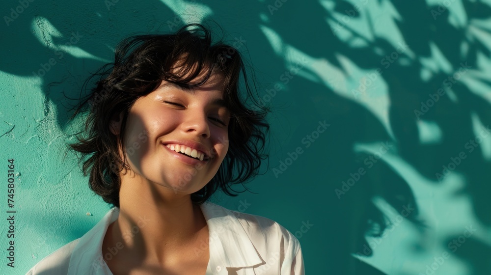 A young woman with dark hair smiling and looking away from the camera with her head resting on a blue wall surrounded by soft blurred shadows of leaves. A young woman with dark hair smiling and looking away from the camera with her head resting on a blue wall surrounded by soft blurred shadows of leaves.