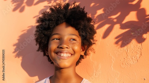 A young child with curly hair smiling brightly standing in front of a textured orange wall with shadows of leaves.