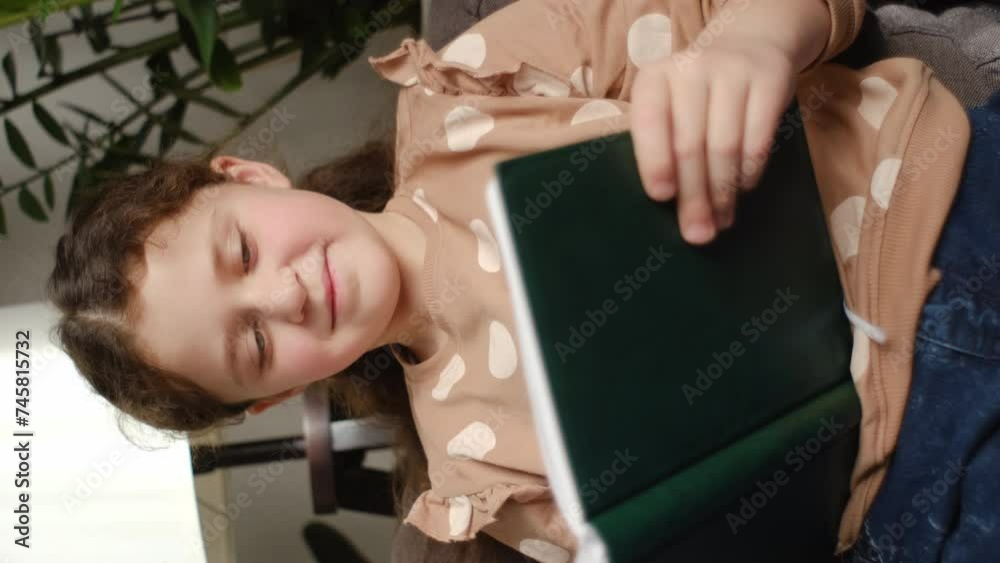 Vertical video of happy cute preteen girl smiling and reading book while sitting on cozy sofa in living room at home. Pretty smiling little child relax reading book at couch. Back to school concept Stock ビデオ | Adobe Stock