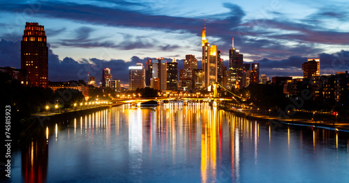 Colorful skyline panorama of the Frankfurt am Main, Germany. Silhouettes of illuminated high-rise office towers on a summer day at blue hour after sunset. Tall buildingss reflected by water surface photo