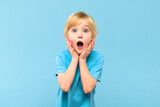 Wow! Portrait of a shocked cute little boy with blond hair on pastel blue background. Surprised preschooler studio shot, looking at camera.