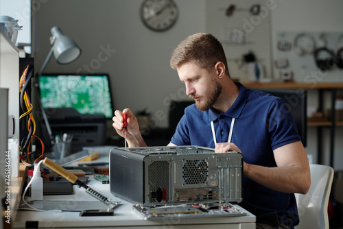 Young serious male technician in dark blue uniform sitting by workplace in repair service office and unfixing parts of computer processor photo